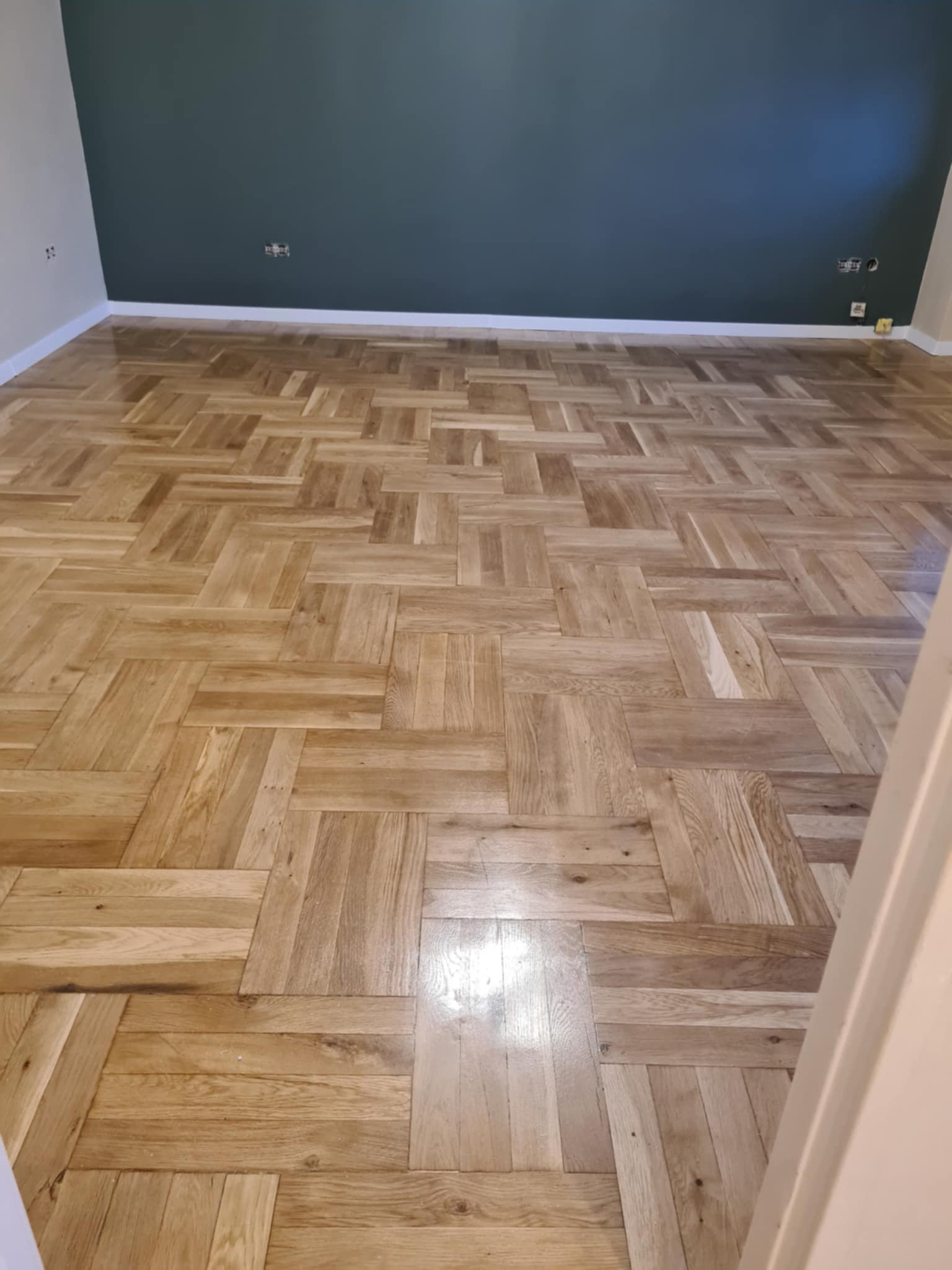 Parkiet ułożony we wzór jodełki francuskiej, po renowacji, w pokoju z ciemnozieloną ścianą, widoczne gniazdka elektryczne.