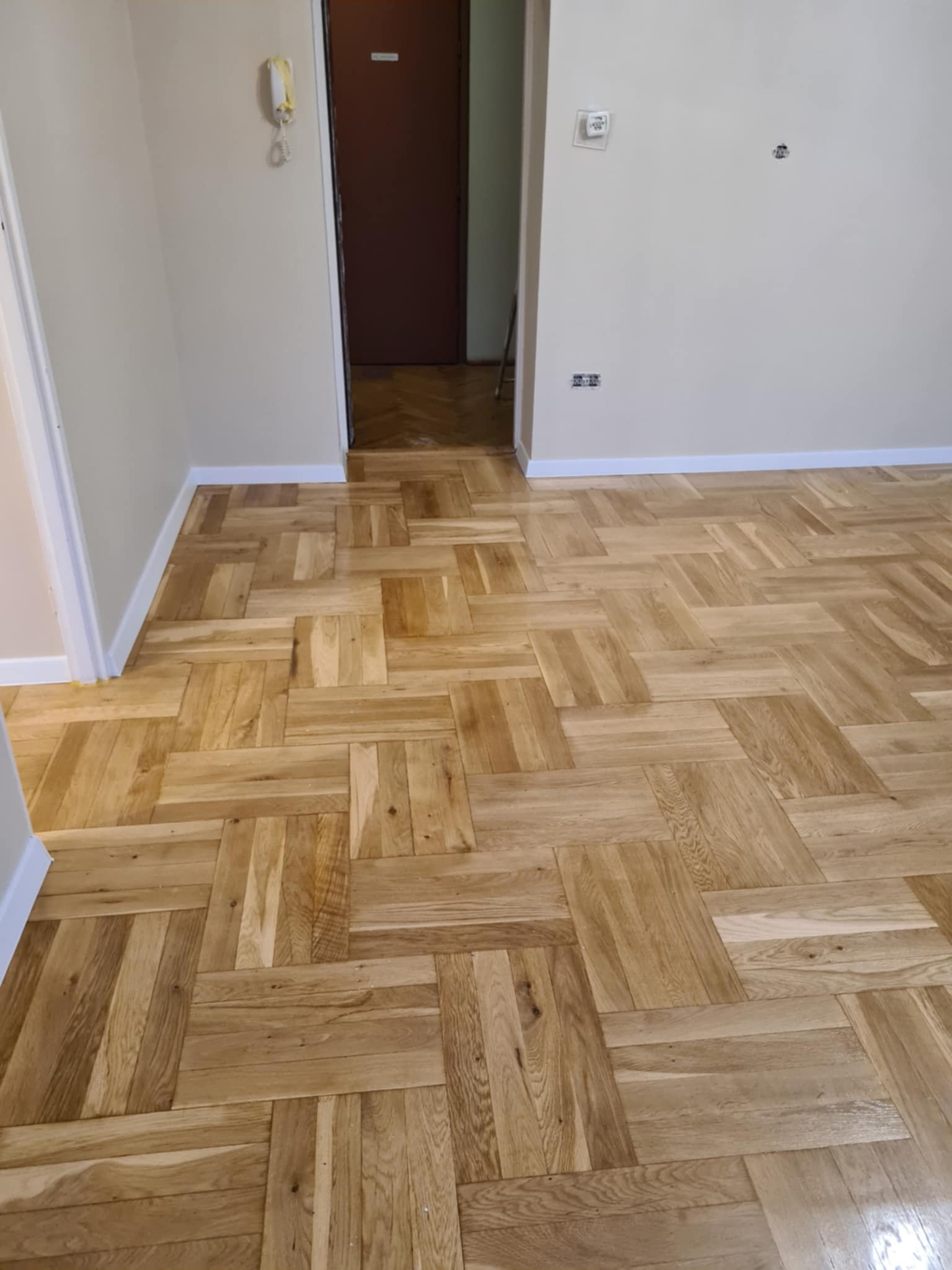 Parkiet ułożony w jodełkę kwadratową, świeżo po cyklinowaniu, widoczne jasne drewno i białe listwy przypodłogowe.