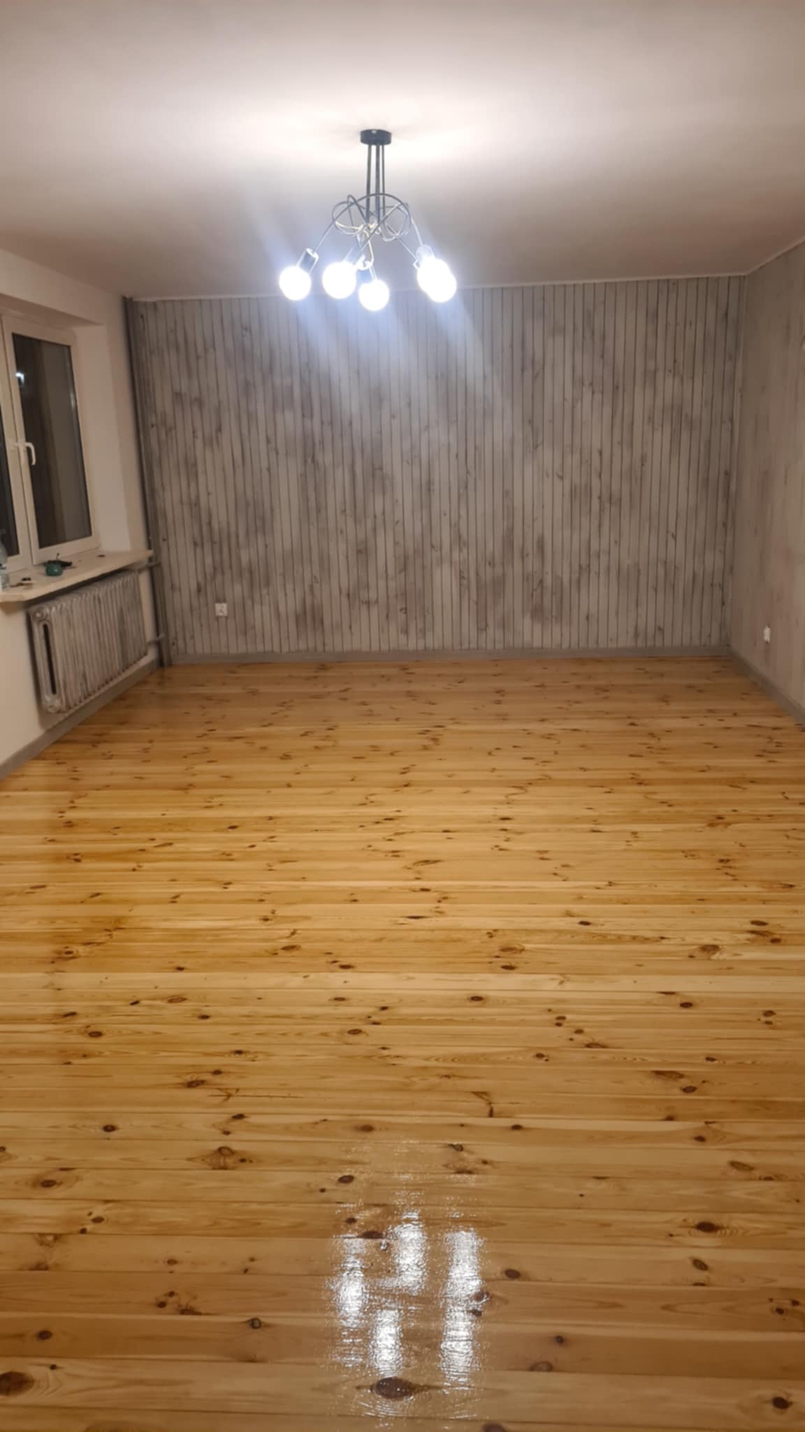 Błyszcząca, świeżo polakierowana podłoga z jasnego drewna w pustym pokoju z szarą ścianą imitującą deski i nowoczesną lampą sufitową.