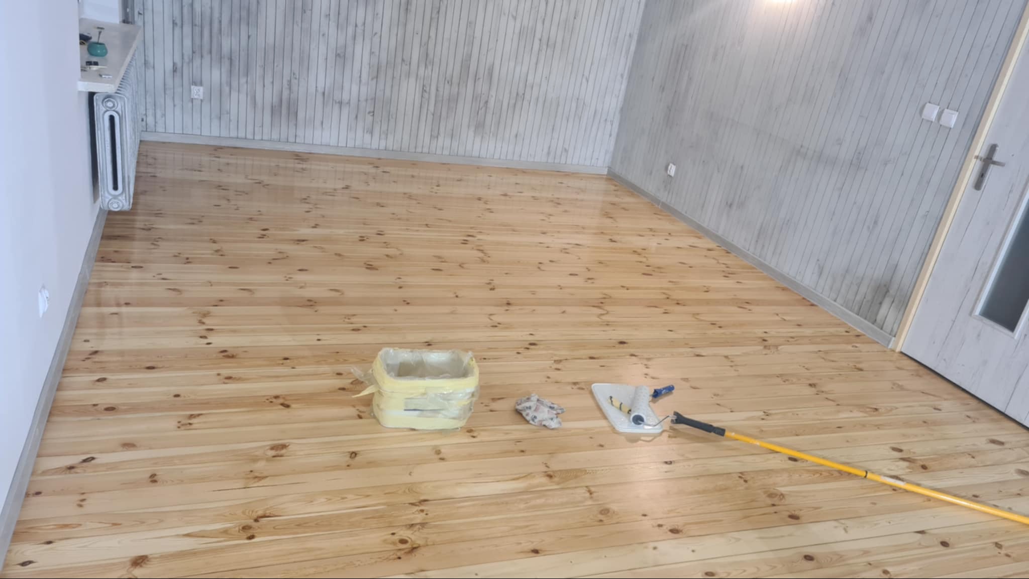 Wycyklinowana podłoga z jasnego drewna, widoczne narzędzia do lakierowania: kuweta z folią, wałek i mop, ściany wykończone boazerią w odcieniach szarości.