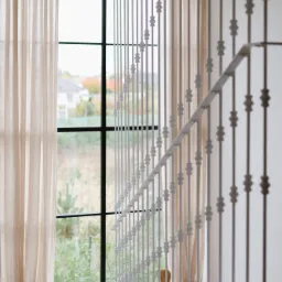 Nowoczesna balustrada schodowa z białych, pionowych prętów z dekoracyjnymi elementami, widok na okno z beżowymi zasłonami i zielonym krajobrazem za oknem.