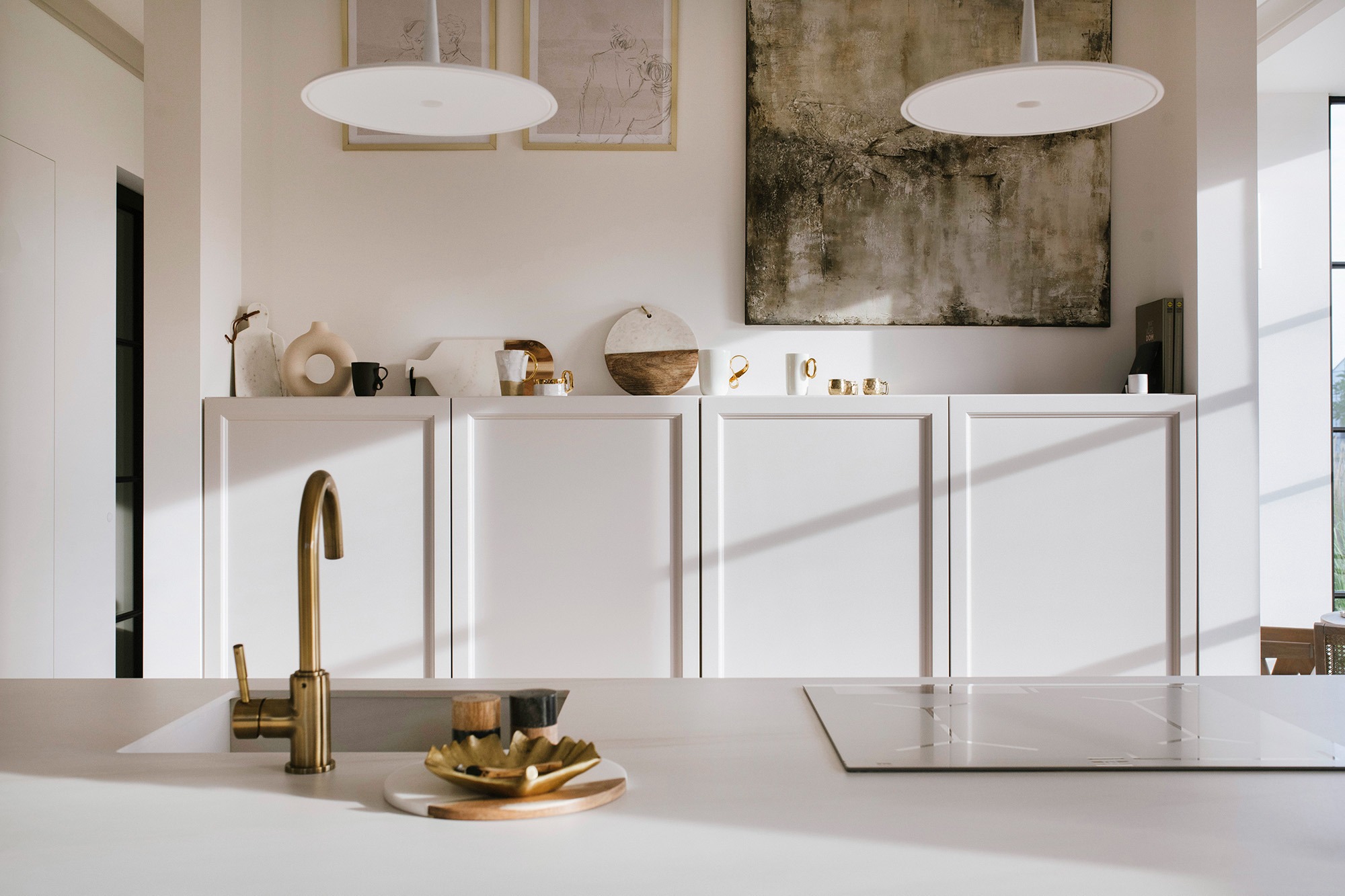 Jasna kuchnia z białymi szafkami, złotą baterią i płytą indukcyjną, dekoracje na blacie i szafkach wiszących, nowoczesne oświetlenie.