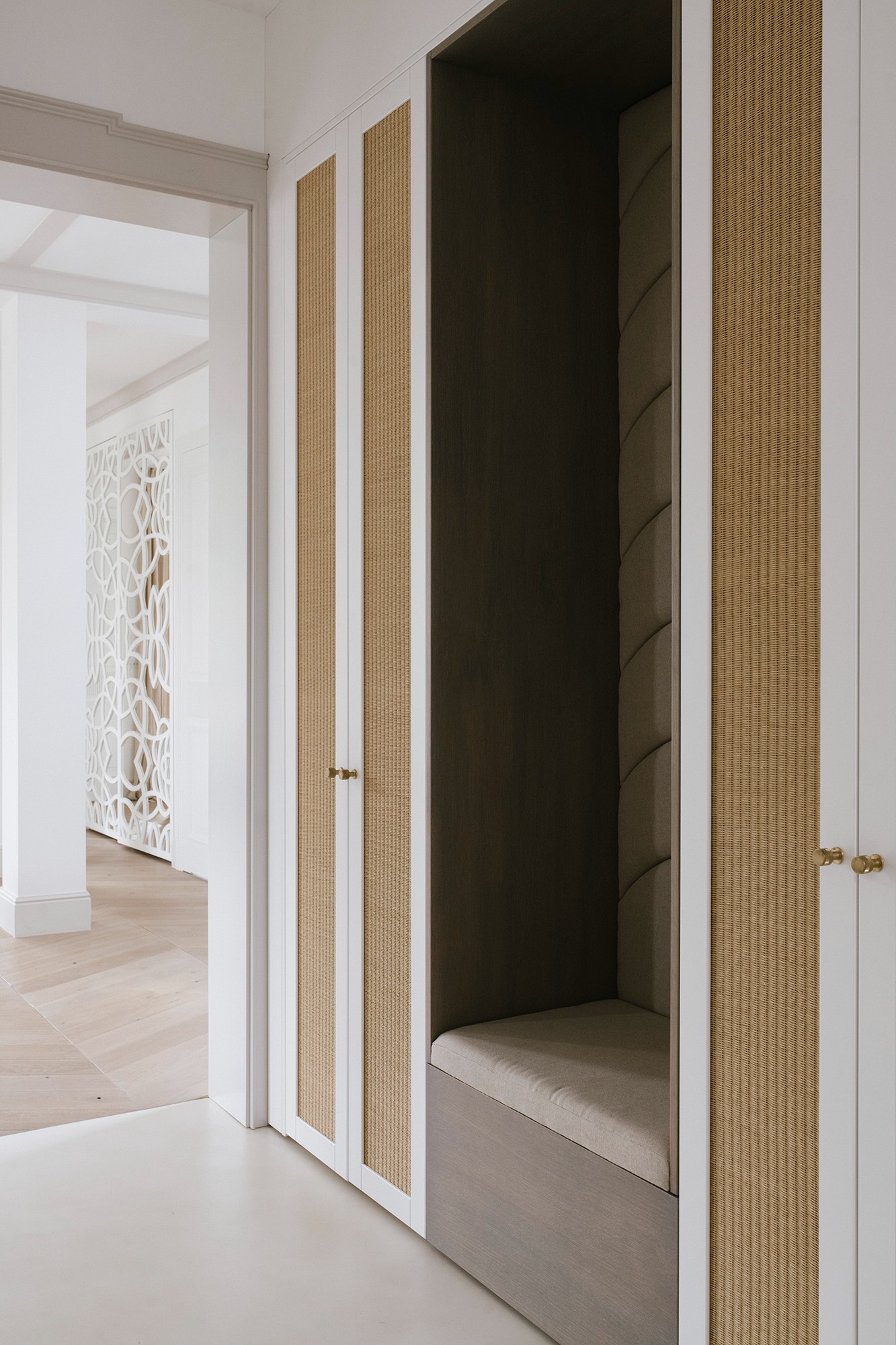 Wnęka z siedziskiem obita tapicerowanym panelem, otoczona szafami z frontami z plecionki w jasnym odcieniu, widok z korytarza z dekoracyjną ścianką ażurową w tle.
