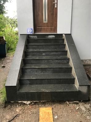 Zewnętrzne schody wejściowe do domu, wyłożone ciemnym, polerowanym granitem, z numerem domu '19' widocznym nad drzwiami.