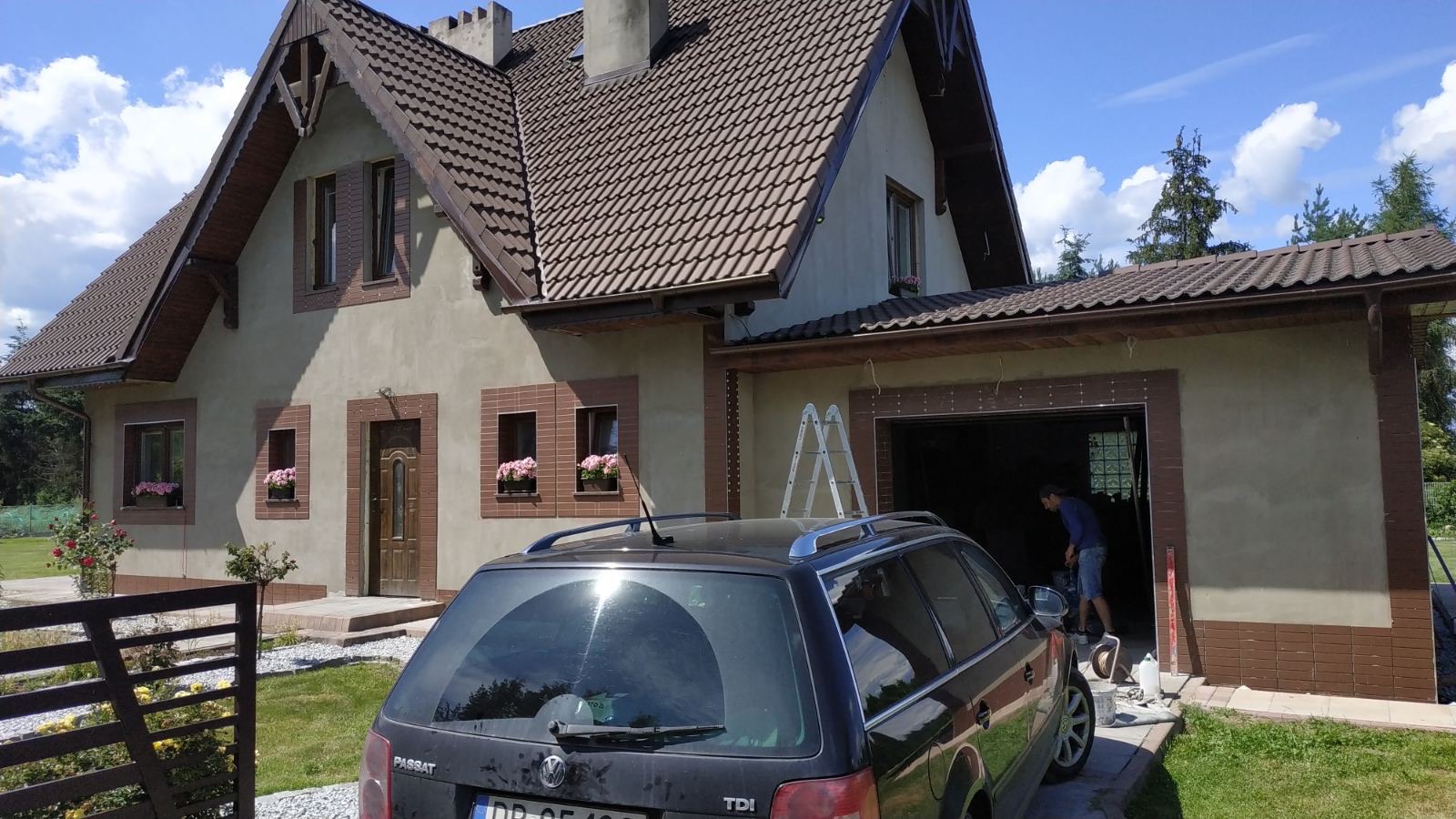 Dwukondygnacyjny dom z brązowym dachem i okiennicami, widoczne kwiaty w oknach, garaż z drabinką i mężczyzną w środku, czarny samochód kombi na pierwszym planie.
