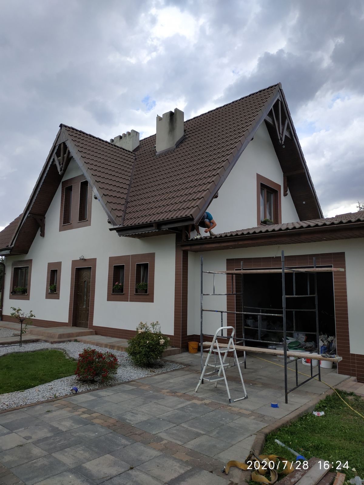 Dwukondygnacyjny dom jednorodzinny z brązowym dachem i białymi ścianami, z pracownikiem na rusztowaniu przy okapie. Widoczne narzędzia i materiały budowlane. Częściowo wykończona elewacja, widoczny...