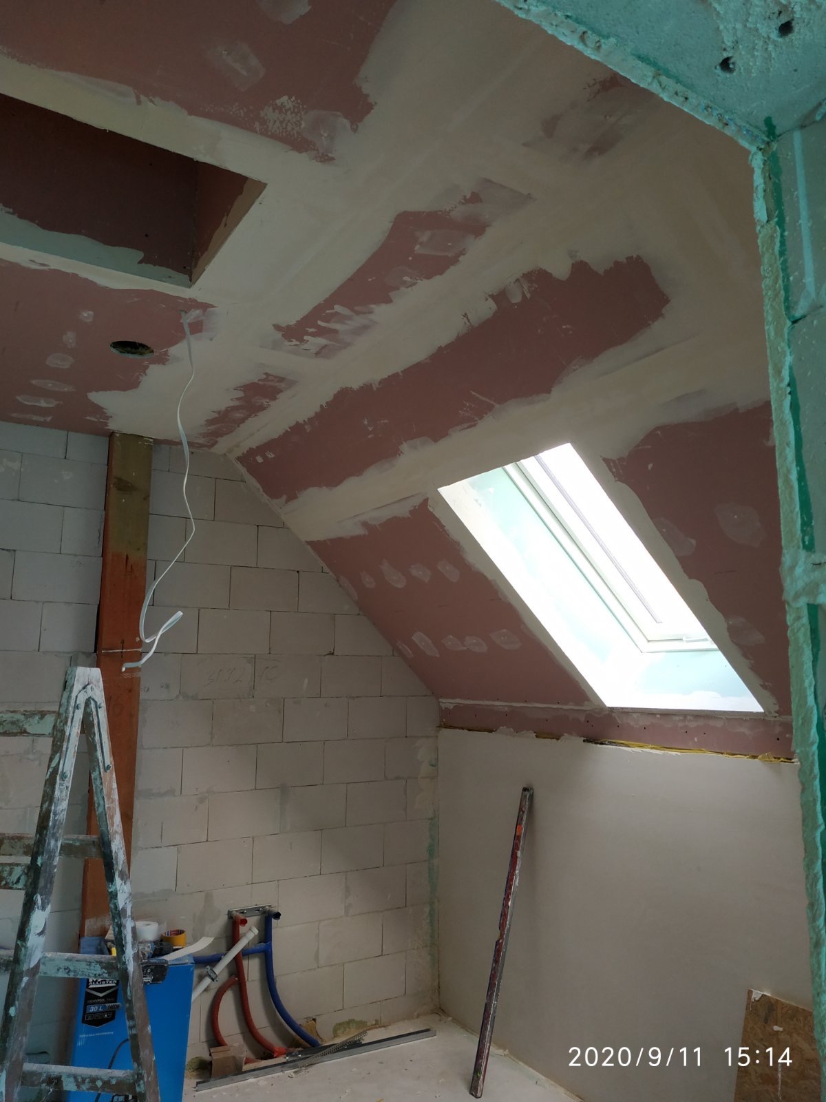 Poddasze w trakcie prac wykończeniowych: ściany z bloczków, sufit z płyt gipsowych z widocznymi łączeniami i oknem dachowym, częściowo nałożona biała gładź, widoczna drabina i narzędzia.