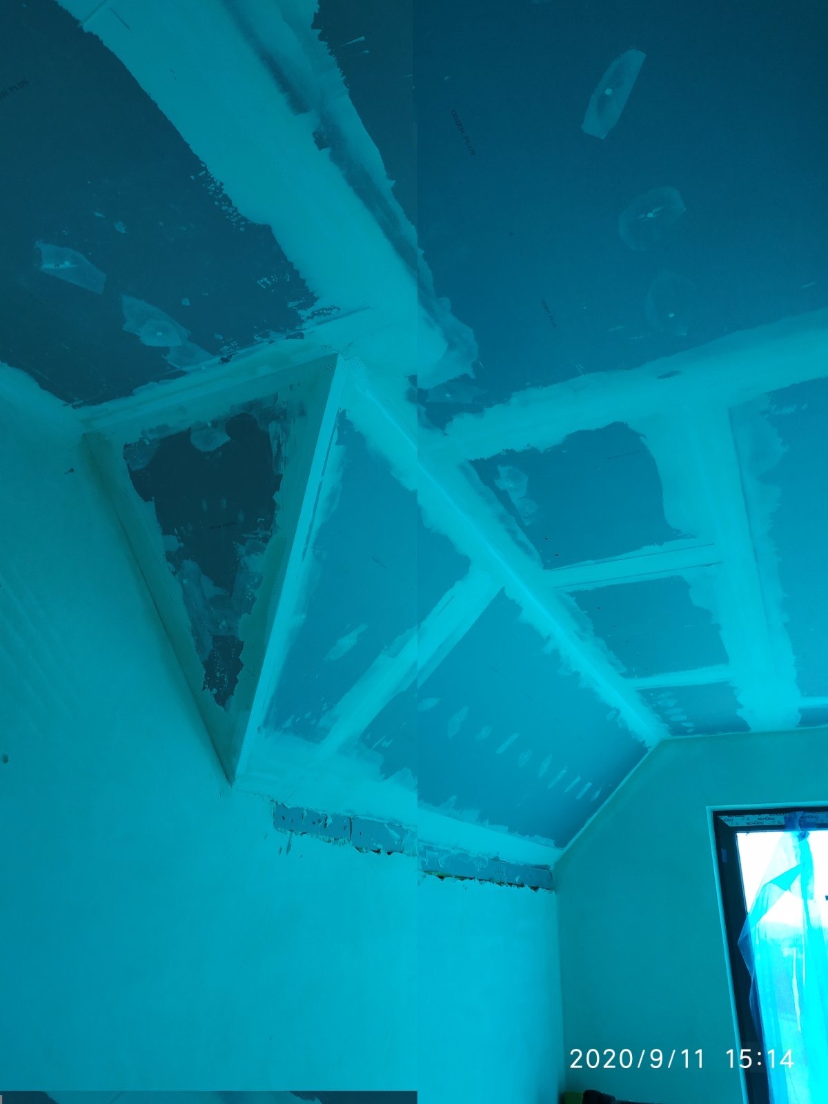 Niedokończona sufitowa konstrukcja z płyt gipsowo-kartonowych, widoczne łączenia i fragmenty niezagruntowanej powierzchni, oświetlenie budowlane w tle.