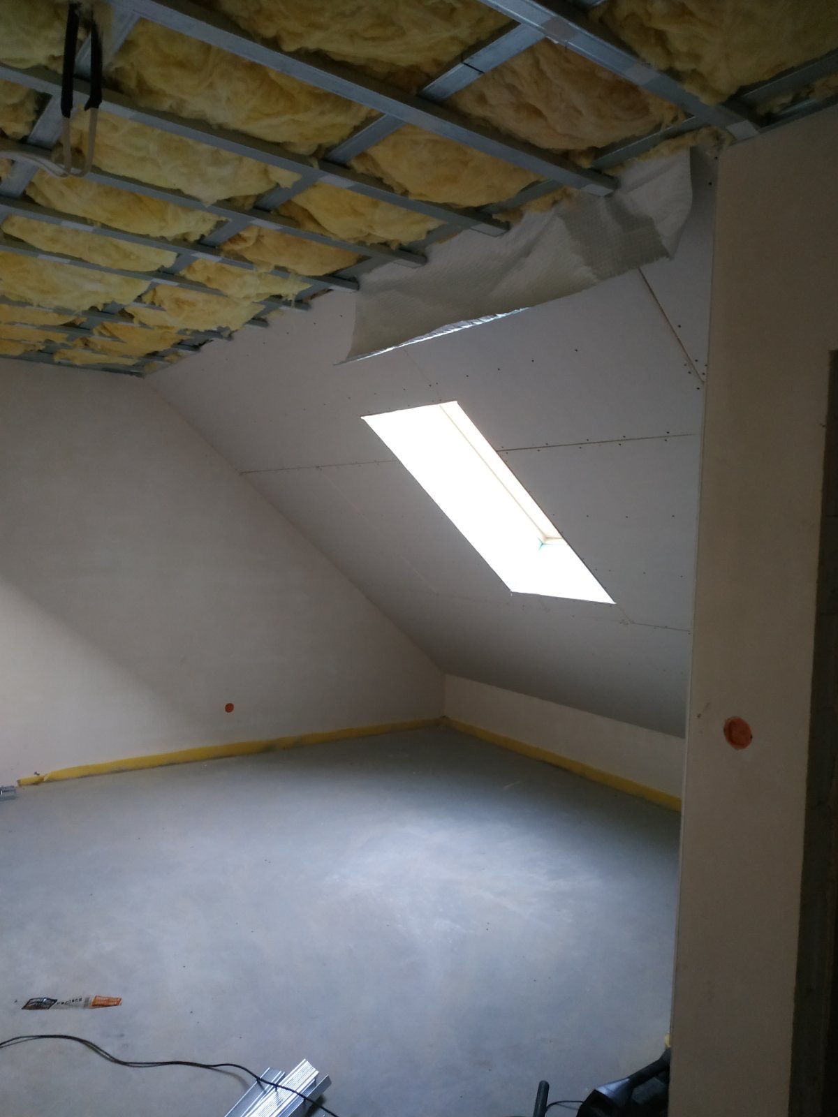 Poddasze w trakcie remontu: widoczna izolacja z wełny mineralnej pod dachem, zamontowane płyty gipsowo-kartonowe ze świetlikiem, surowa wylewka na podłodze i zabezpieczona folią ściana.