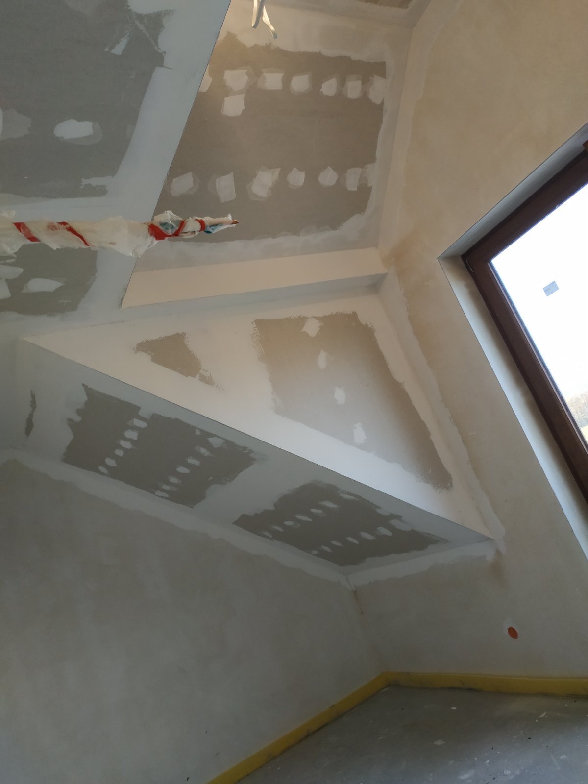 Wnętrze w trakcie prac przygotowawczych do wykończenia, ściany i sufit z widocznymi łatami gipsowymi, zabezpieczona folią rura, okno z drewnianą ramą.