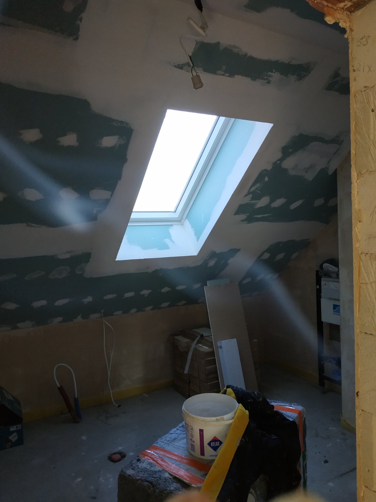 Poddasze w trakcie prac wykończeniowych: widoczne płyty gipsowo-kartonowe na skosach, szpachlowane łączenia, okno dachowe, wiadro z zaprawą Atlas i inne materiały budowlane.