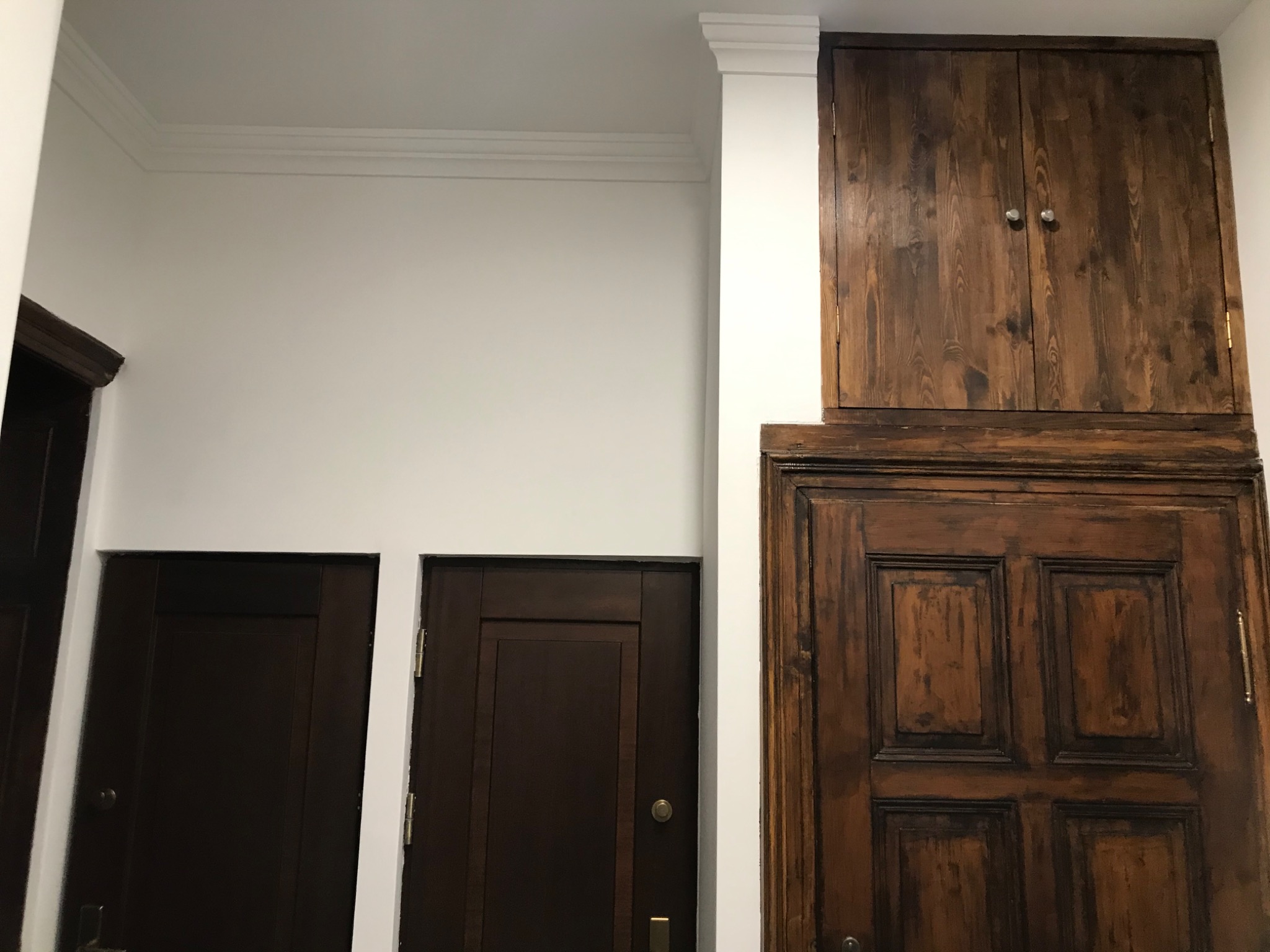Dwa ciemnobrązowe drzwi wewnętrzne obok siebie, jedno z nich z dodatkową szafką nadprożową z drewna o widocznych słojach, białe ściany i listwy przypodłogowe.