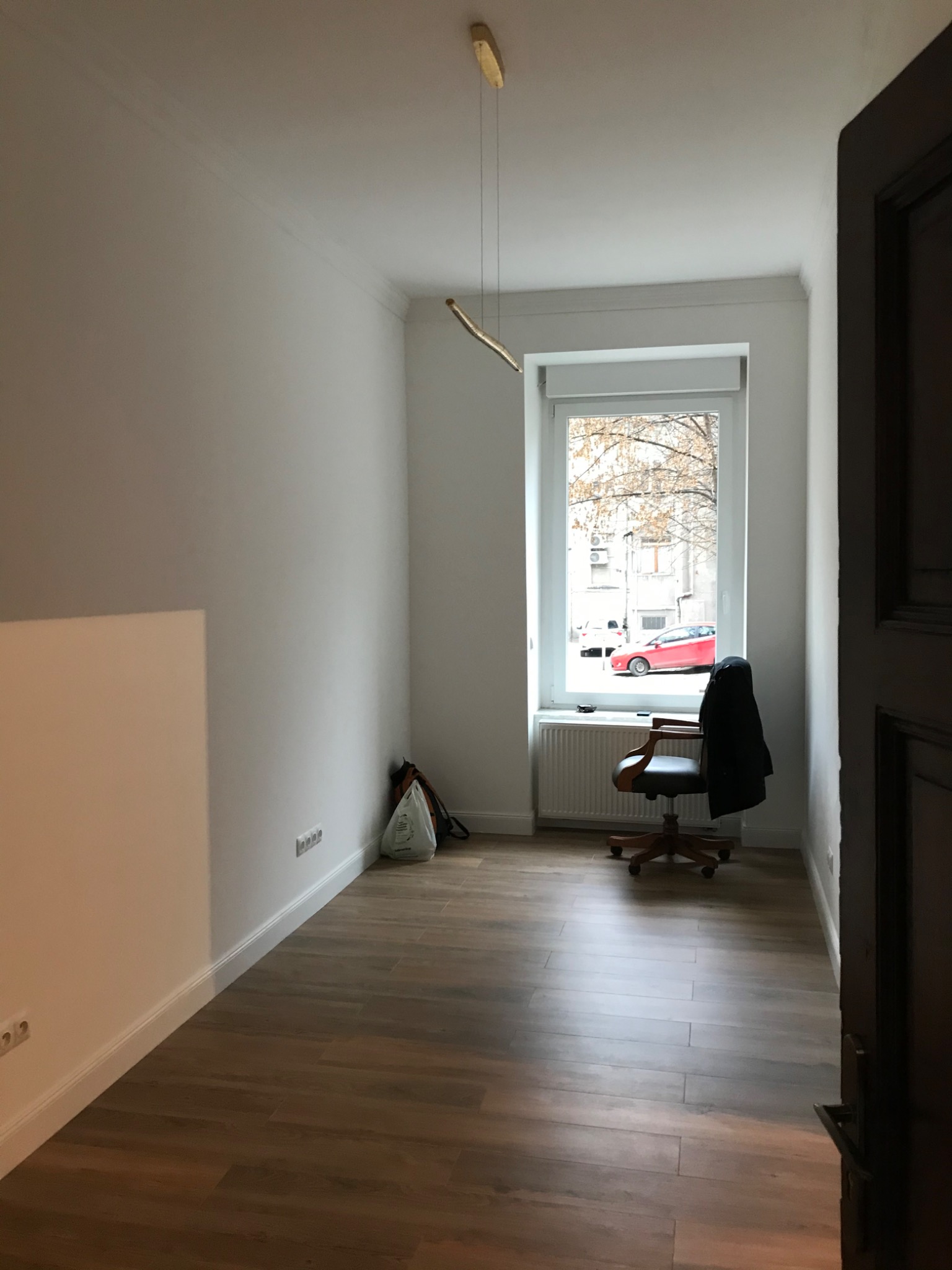 Pusty pokój po remoncie z drewnianą podłogą, białymi ścianami i nowoczesną lampą wiszącą, widok zza otwartych drzwi.