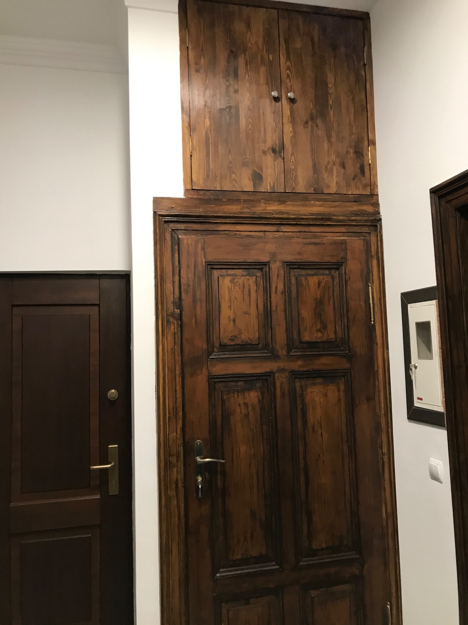 Ciemne, drewniane drzwi wewnętrzne z górną szafką w stylu retro, widoczne obok brązowych drzwi i skrzynki elektrycznej na białej ścianie.