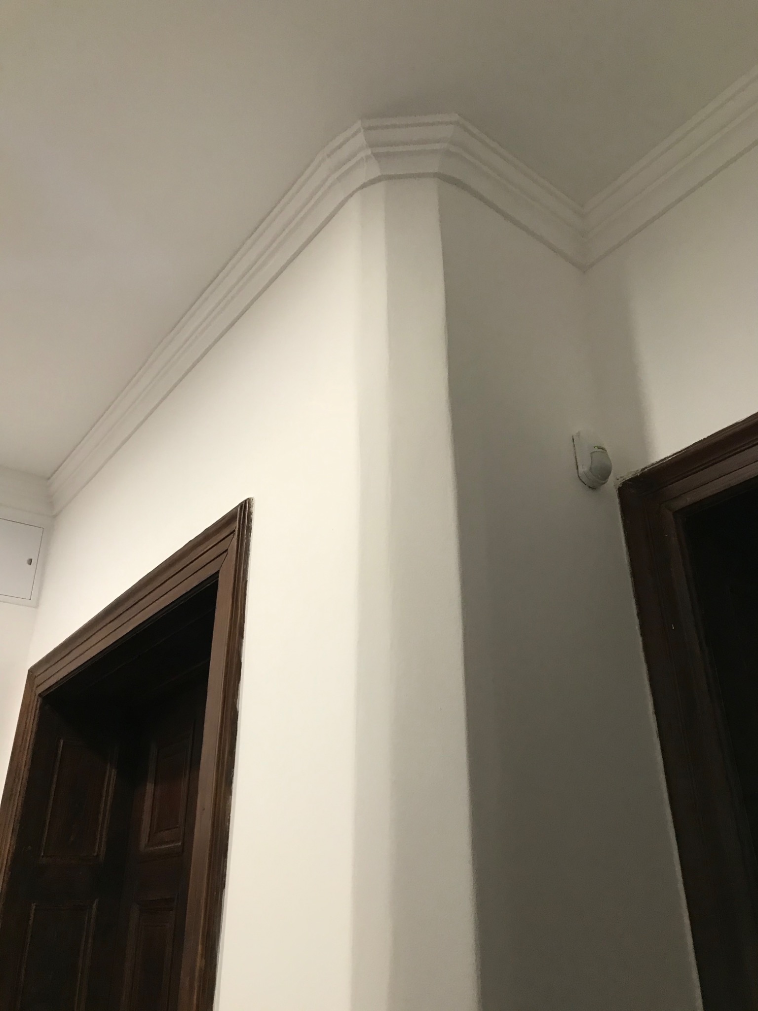 Pomieszczenie po remoncie: gładkie, białe ściany, ozdobny gzyms sufitowy i ciemne, drewniane ościeżnice drzwiowe.