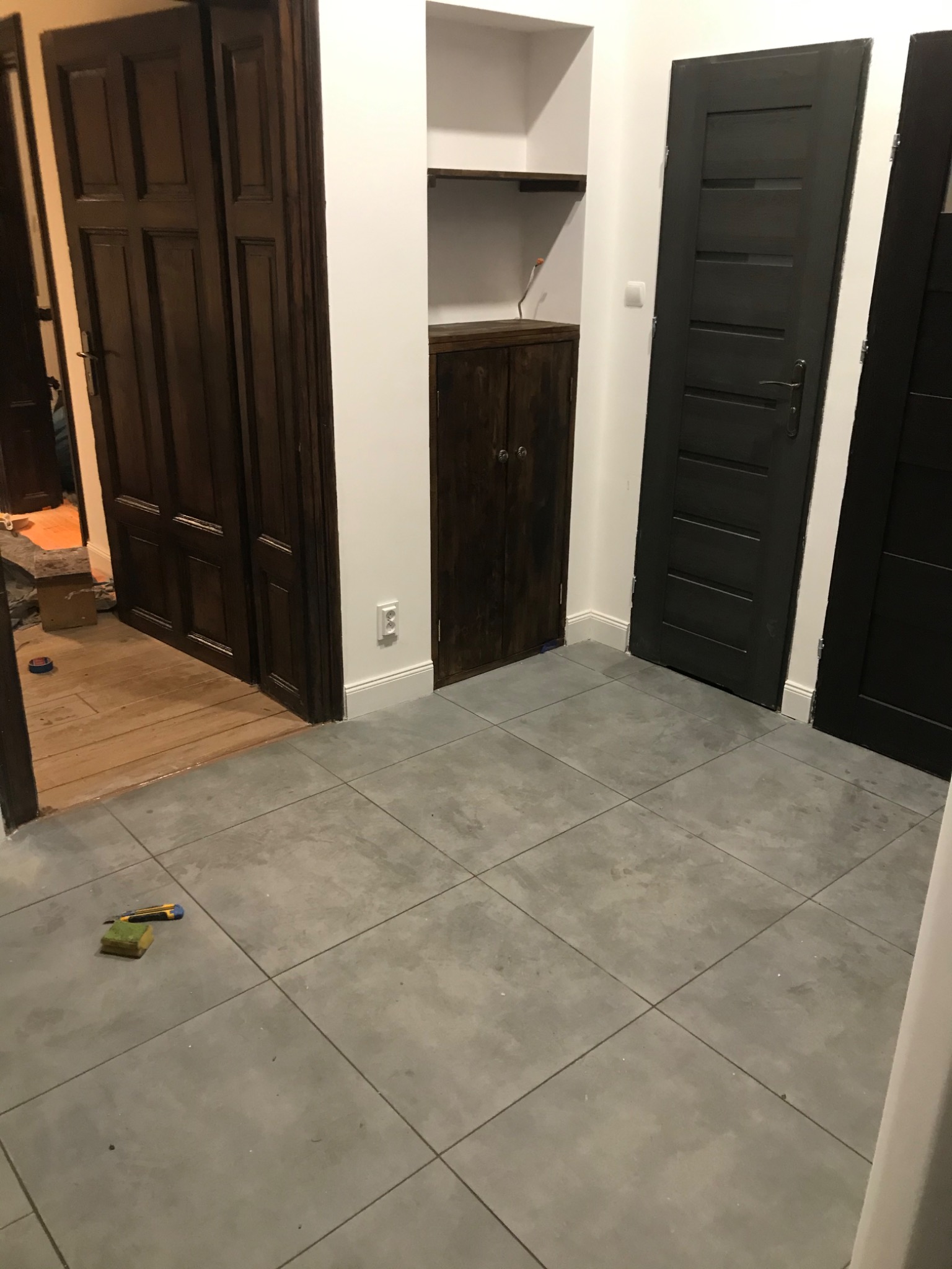 Wnętrze mieszkania w trakcie remontu, widoczne dwie pary drzwi w różnych kolorach, szare płytki na podłodze i narzędzia do cięcia dywanów.