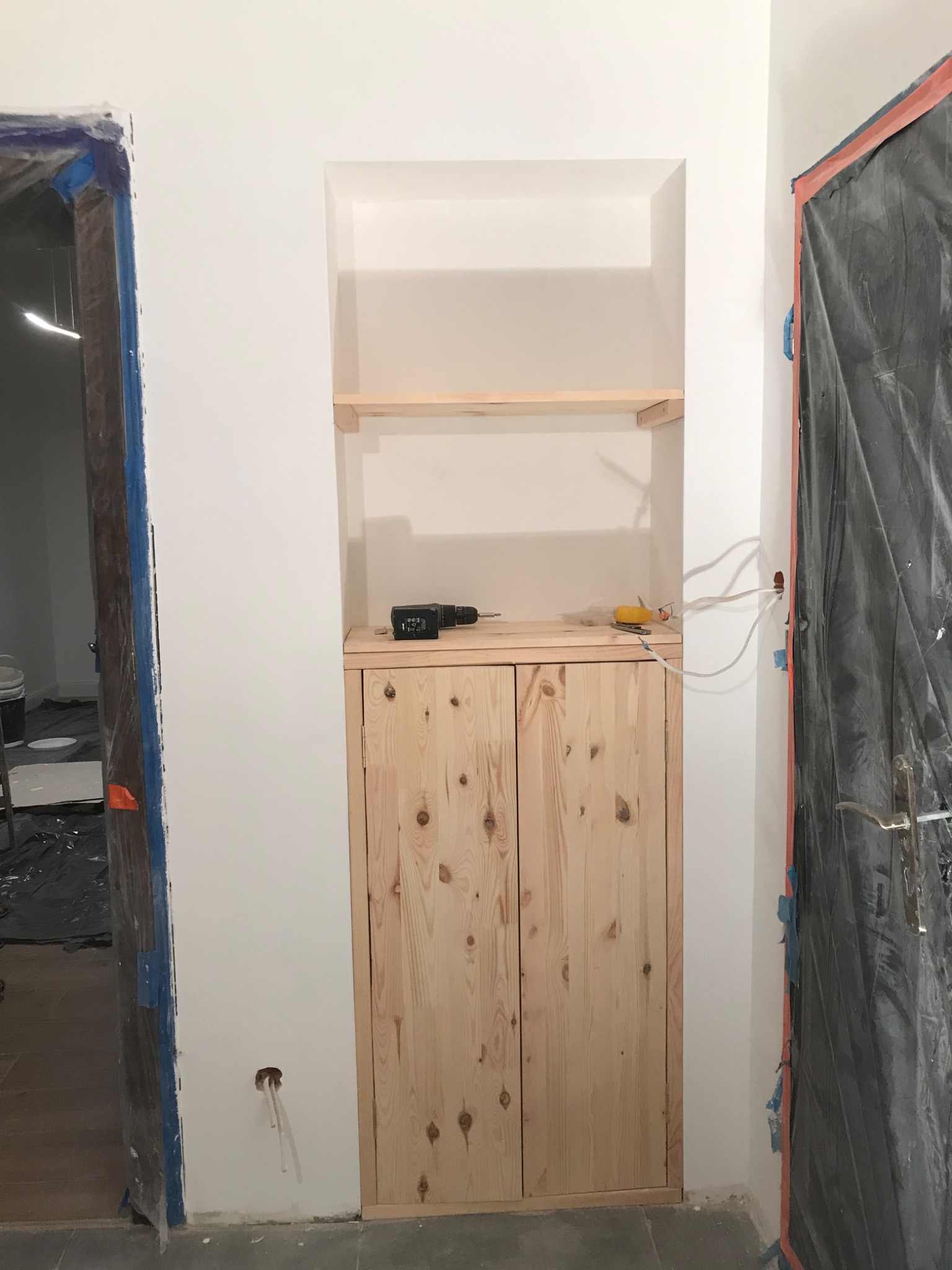 Wnęka z drewnianymi półkami i szafką podczas prac wykończeniowych we Wrocławiu, widoczne zabezpieczone folią drzwi, narzędzia na szafce i wystające przewody.