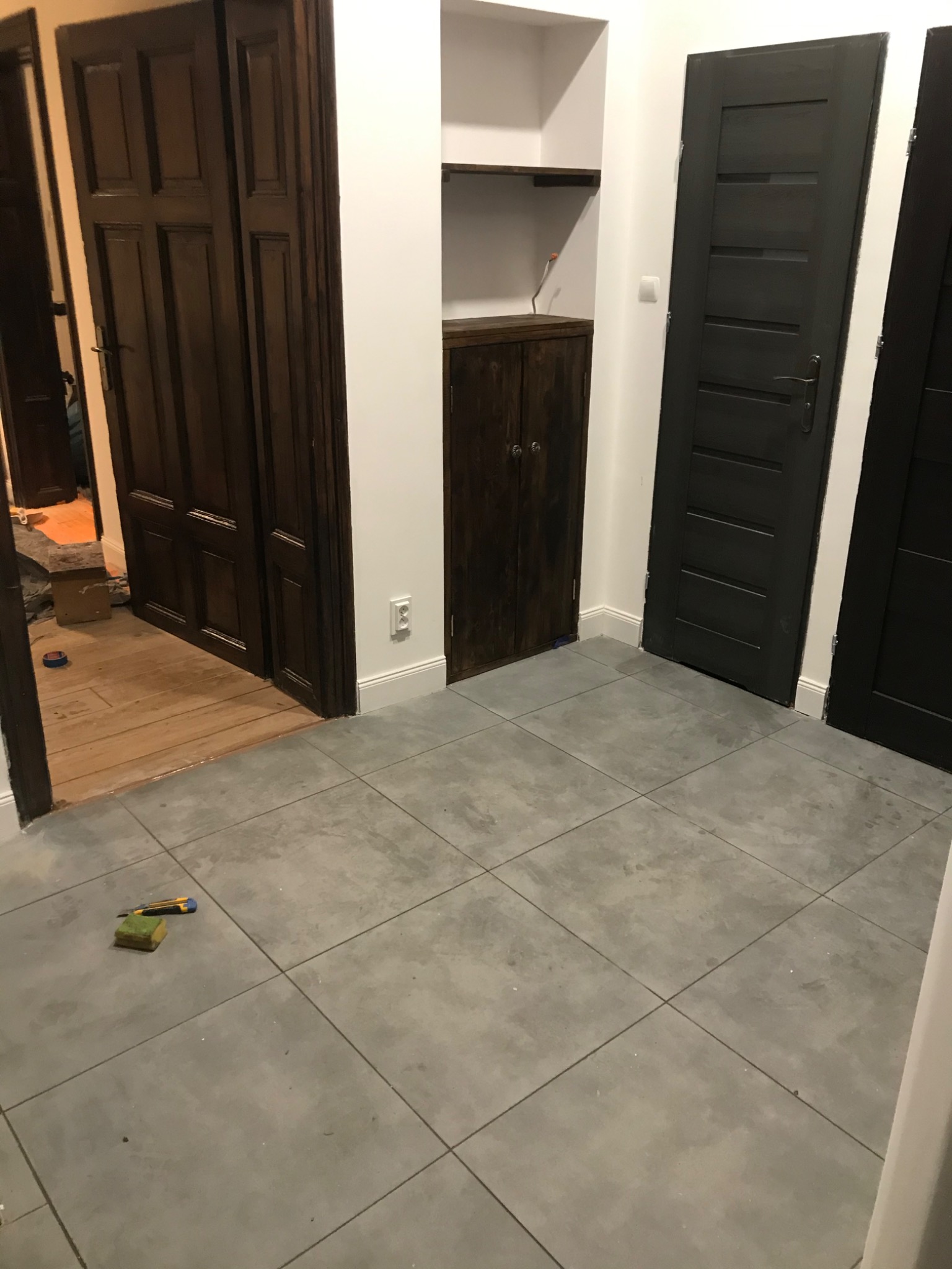 Korytarz w trakcie remontu z szarymi płytkami podłogowymi, widoczne dwie pary ciemnych drewnianych drzwi, szafka wnękowa, narzędzia do cięcia i gąbka na podłodze, w tle fragment podłogi wyłożonej...