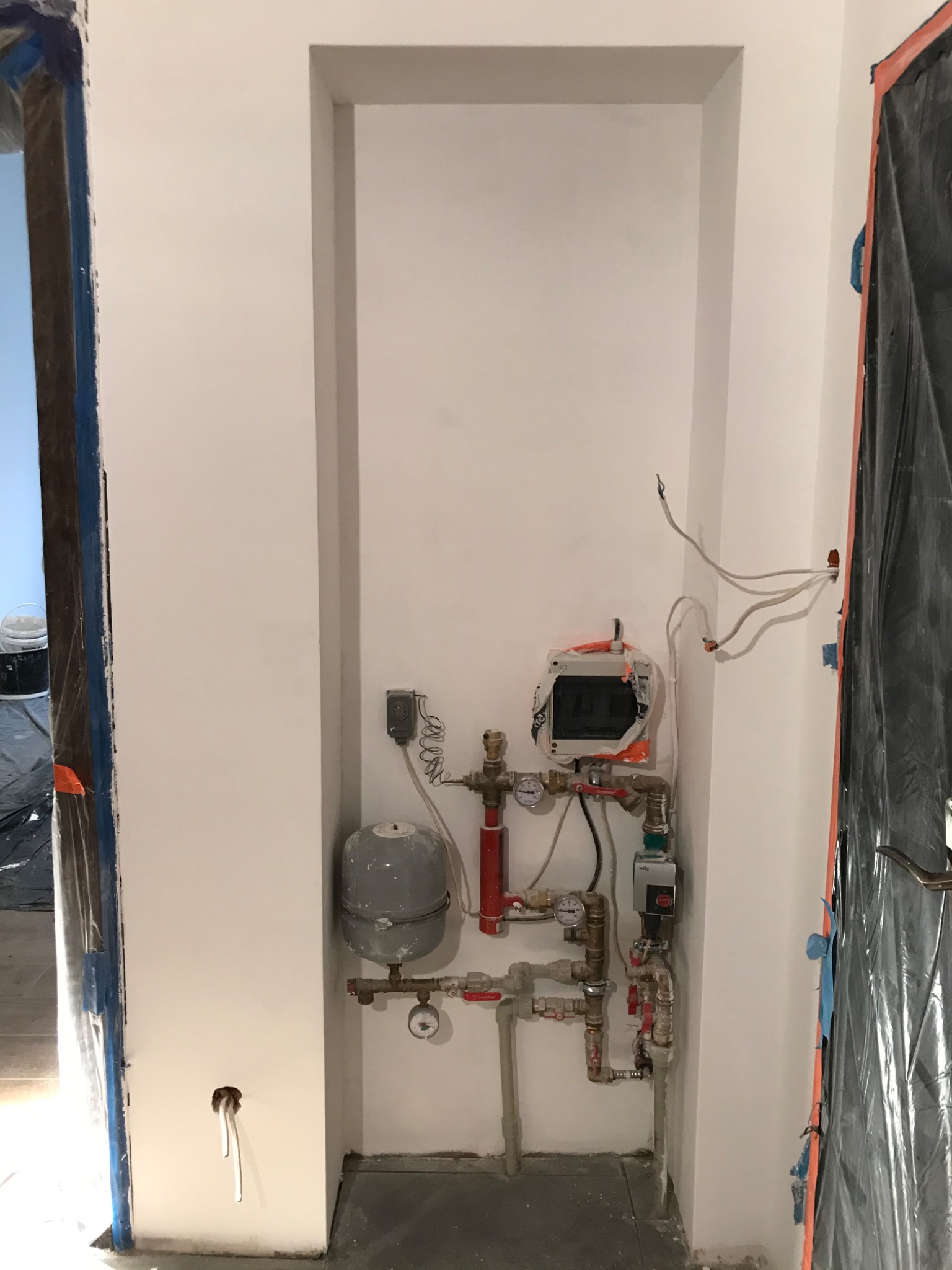 Wnęka w ścianie z instalacją centralnego ogrzewania w trakcie montażu, widoczne przewody, rury, manometry, zabezpieczona folią futryna drzwi.