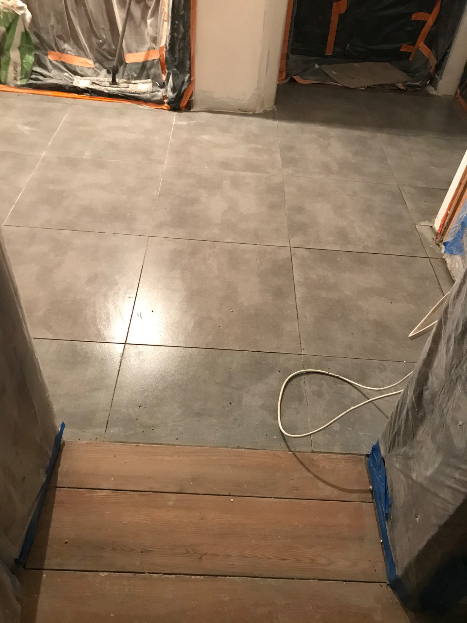 Ułożone szare płytki podłogowe w trakcie remontu, zabezpieczone folią drzwi i ściany, widoczny kabel elektryczny na podłodze.