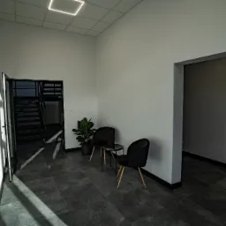 Nowoczesna przestrzeń biurowa z dwoma czarnymi krzesłami przy małym stoliku, roślina doniczkowa, widok na schody i biuro, minimalistyczny design, gra świateł słonecznych na podłodze.