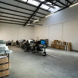 Przestronne biuro z pracownikami przy komputerach, stertami dokumentów i kartonami, oświetlone naturalnym światłem z okien dachowych.