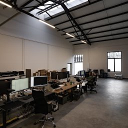 Archiveo Sp. ZO. O - Przestronne biuro z wysokim sufitem i belkami, wyposażone w stanowiska pracy z monitorami i skanerami, kartony i segregatory z dokumentami, widoczne okno i oświetlenie sufitowe.