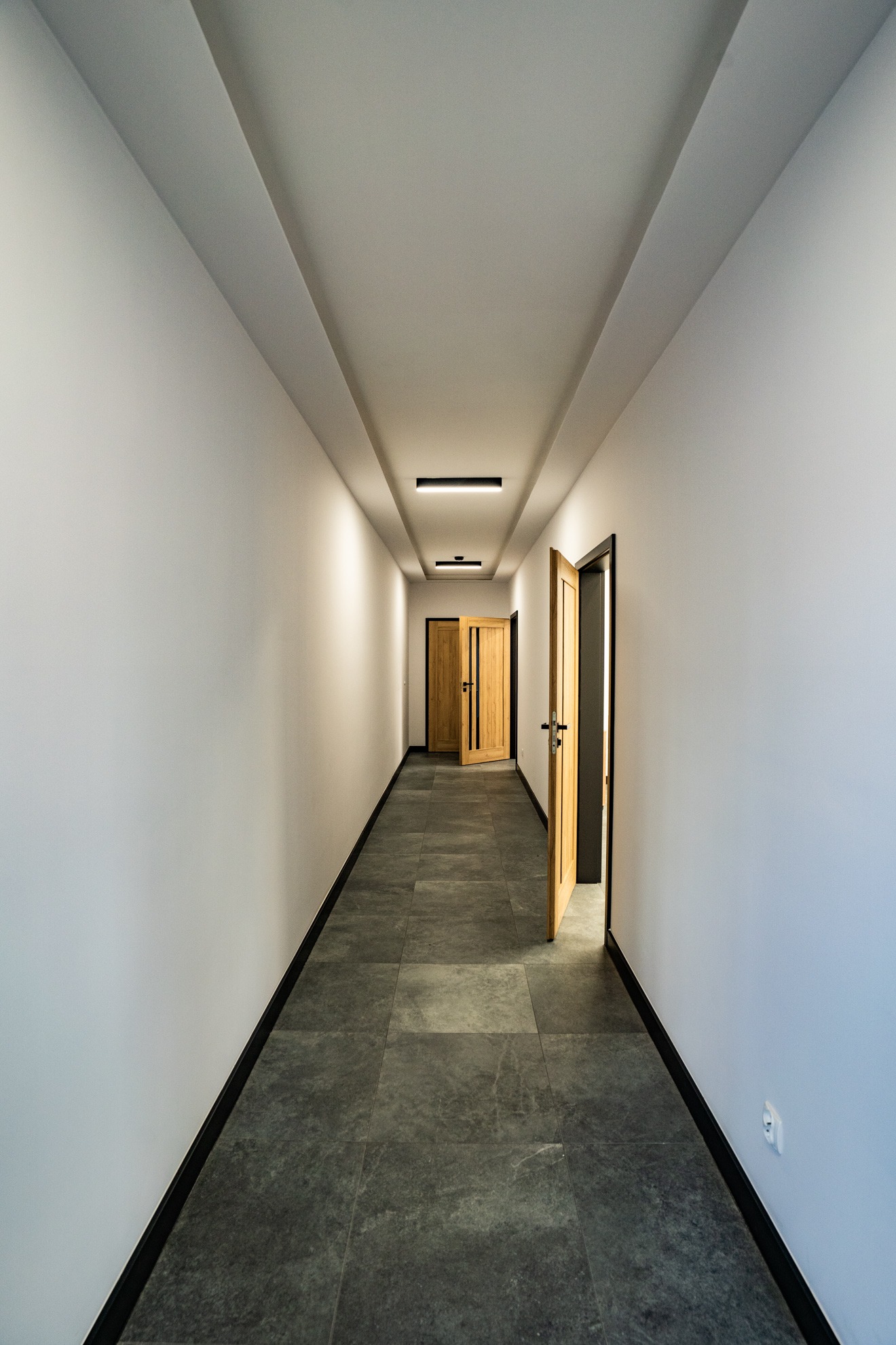 Długi, nowoczesny korytarz biurowy z szarymi płytkami podłogowymi, białymi ścianami, czarnymi listwami przypodłogowymi i drewnianymi drzwiami, z których jedne są otwarte.