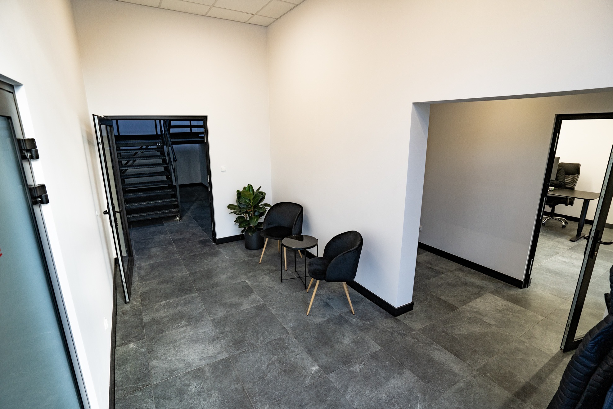 Wnętrze biura z dwoma czarnymi krzesłami i małym stolikiem, widok na schody i biuro z biurkiem, szara podłoga.