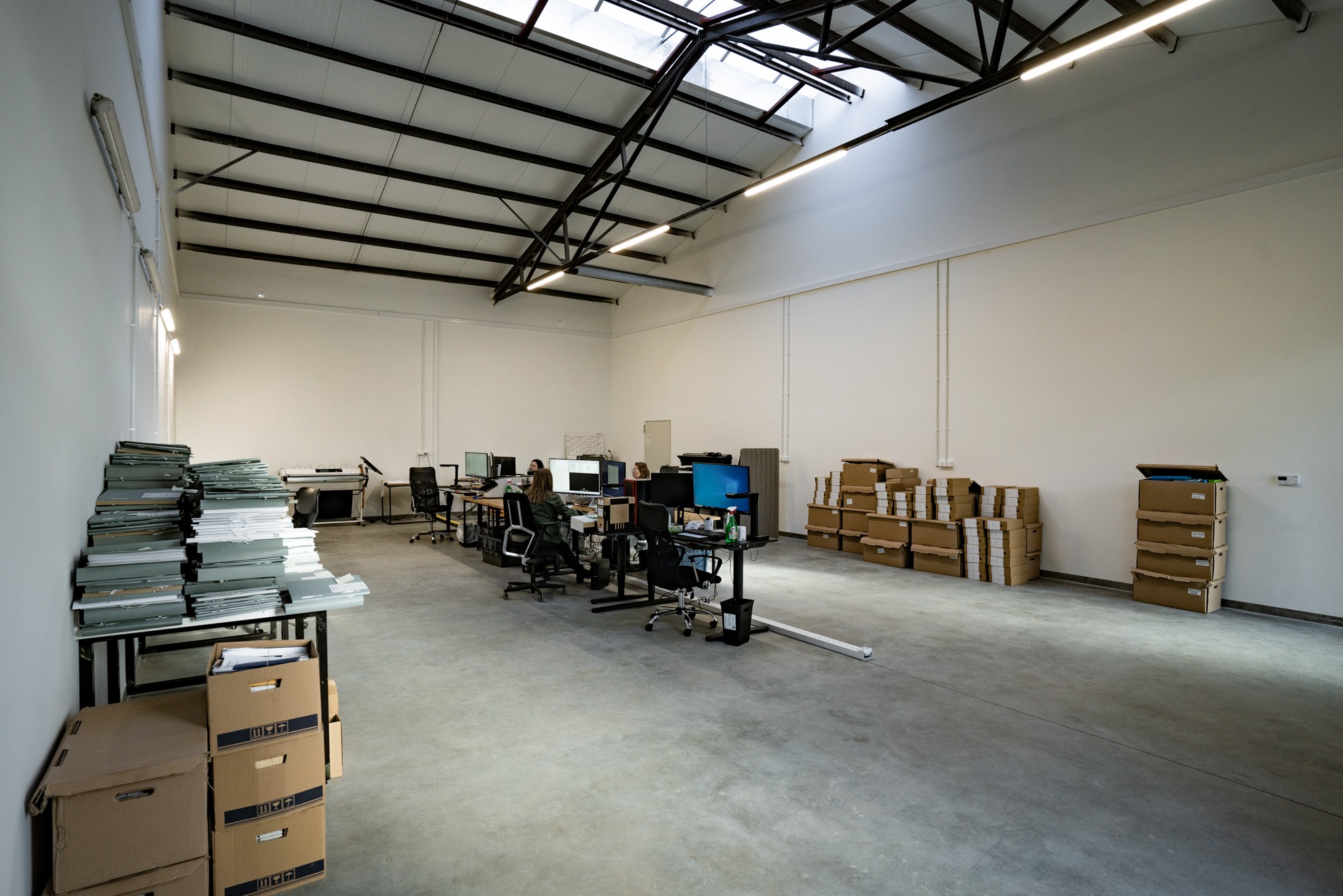 Przestronne biuro z pracownikami przy komputerach, stertami dokumentów i kartonami, oświetlone naturalnym światłem z okien dachowych.