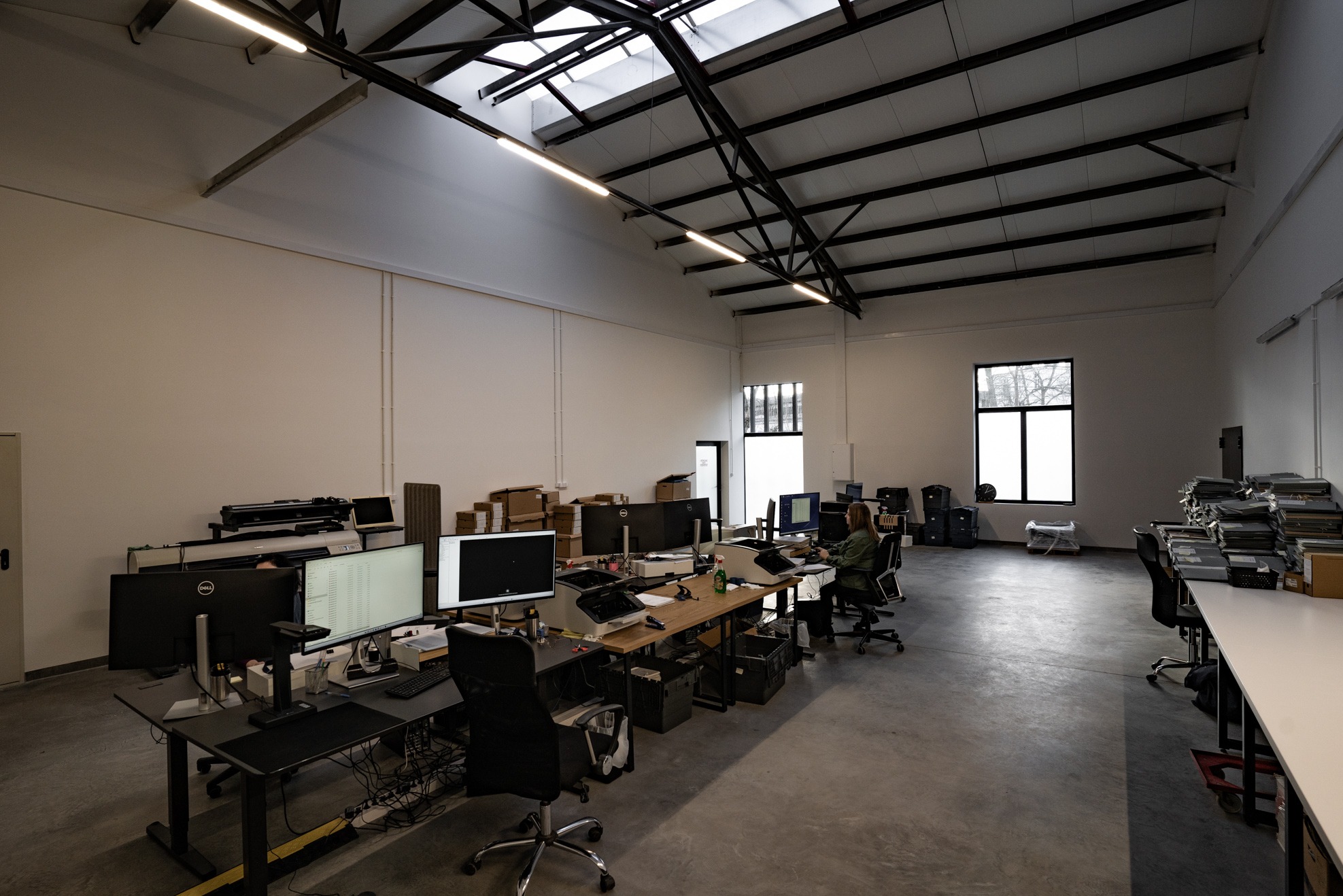 Przestronne biuro z wysokim sufitem i belkami, wyposażone w stanowiska pracy z monitorami i skanerami, kartony i segregatory z dokumentami, widoczne okno i oświetlenie sufitowe.