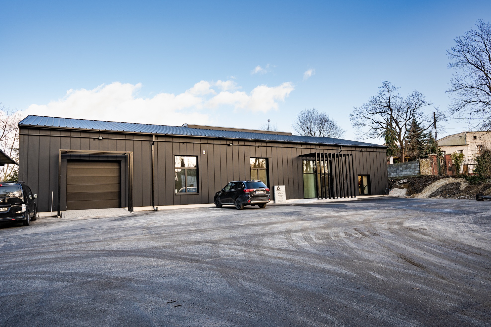 Nowoczesny budynek archiwum z ciemną elewacją i bramą garażową, zaparkowany czarny samochód osobowy, widoczny znak informacyjny.