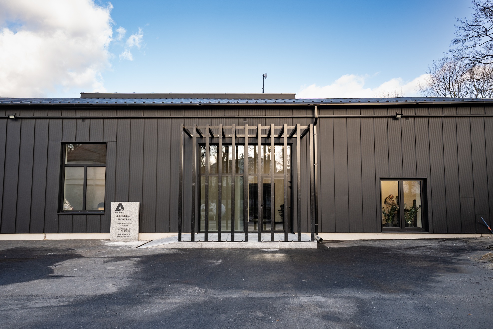 Nowoczesny budynek biurowy w ciemnej kolorystyce z elementami dekoracyjnymi przy wejściu i tablicą informacyjną firmy ArchiVeo. Widoczne okna z roślinami wewnątrz.