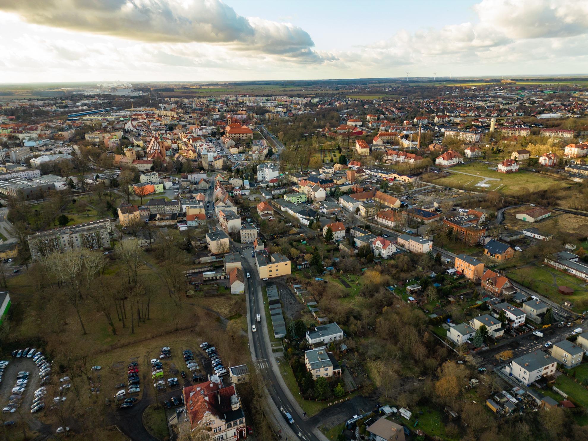 Szeroki widok z lotu ptaka na miasto z budynkami mieszkalnymi, kościołem z czerwoną wieżą, drzewami bez liści i parkingiem pełnym samochodów.