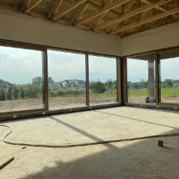 Wnętrze surowego domu w trakcie wykończenia z zamontowanymi dużymi oknami przesuwnymi z brązowymi ramami, widok na zewnątrz na domy i zieleń, na podłodze leży kabel elektryczny i stoi wiadro.