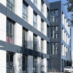 Nowoczesny budynek biurowy z szarymi i białymi panelami elewacyjnymi, liczne okna, rowery i samoch&oacute;d zaparkowane przed budynkiem, pomarańczowe pachołki na chodniku.