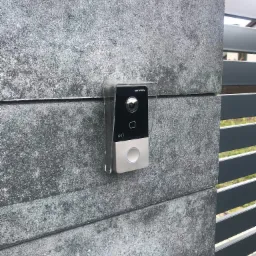 Nowoczesny wideodomofon Hikvision zamontowany na szarym, betonowym słupku ogrodzeniowym, chroniony przez przezroczystą osłonę przeciwdeszczową.