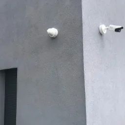 Dwie kamery monitoringu, kopułkowa i tubowa, zamontowane na szarej, teksturowanej elewacji budynku z wąskimi, pionowymi oknami z żaluzjami.