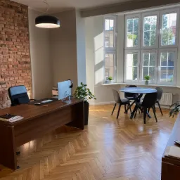 Nowoczesne biuro z ceglaną ścianą i parkietem w jodełkę, wyposażone w drewniane biurko z komputerem, czarne krzesło biurowe, okrągły stół z krzesłami i duże okno z widokiem na zieleń.