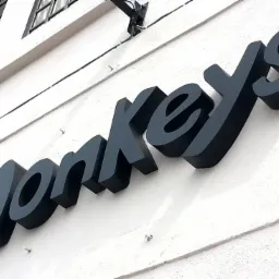 Elewacja budynku z zamontowanym, przestrzennym napisem 'monkeys' wykonanym z ciemnego tworzywa sztucznego, oświetlonym z góry.