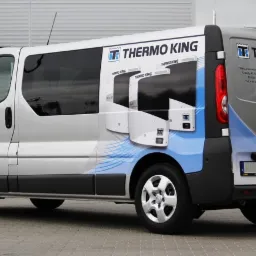 Srebrny van z grafiką reklamową chłodni Thermo King, zaparkowany na zewnątrz budynku, z widocznymi elementami identyfikacji wizualnej firmy.