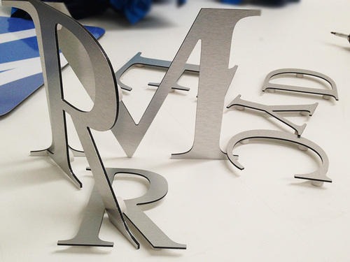 Metalowe litery o szczotkowanej powierzchni, stojące na białym tle, częściowo ułożone w słowo 'MARCYD', z widocznymi detalami wykończenia krawędzi.