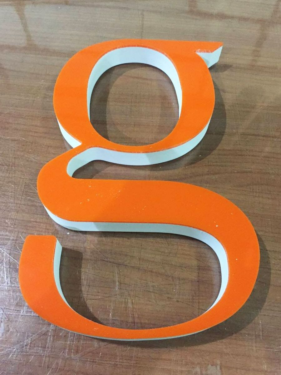 Pomarańczowa litera 'g' wykonana z tworzywa sztucznego z białą krawędzią, leżąca na drewnianej powierzchni z widocznymi rysami.