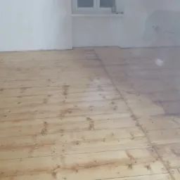 Wypolerowana podłoga z desek w jasnym odcieniu, widoczne ślady po szpachlowaniu i gwoździach, świeżo po nałożeniu lakieru, ściany w tle przygotowane do malowania.