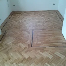 Nowa, jasna podłoga parkietowa ułożona we wzór jodełki francuskiej z ciemną bordiurą w pomieszczeniu z białymi ścianami i oknem.