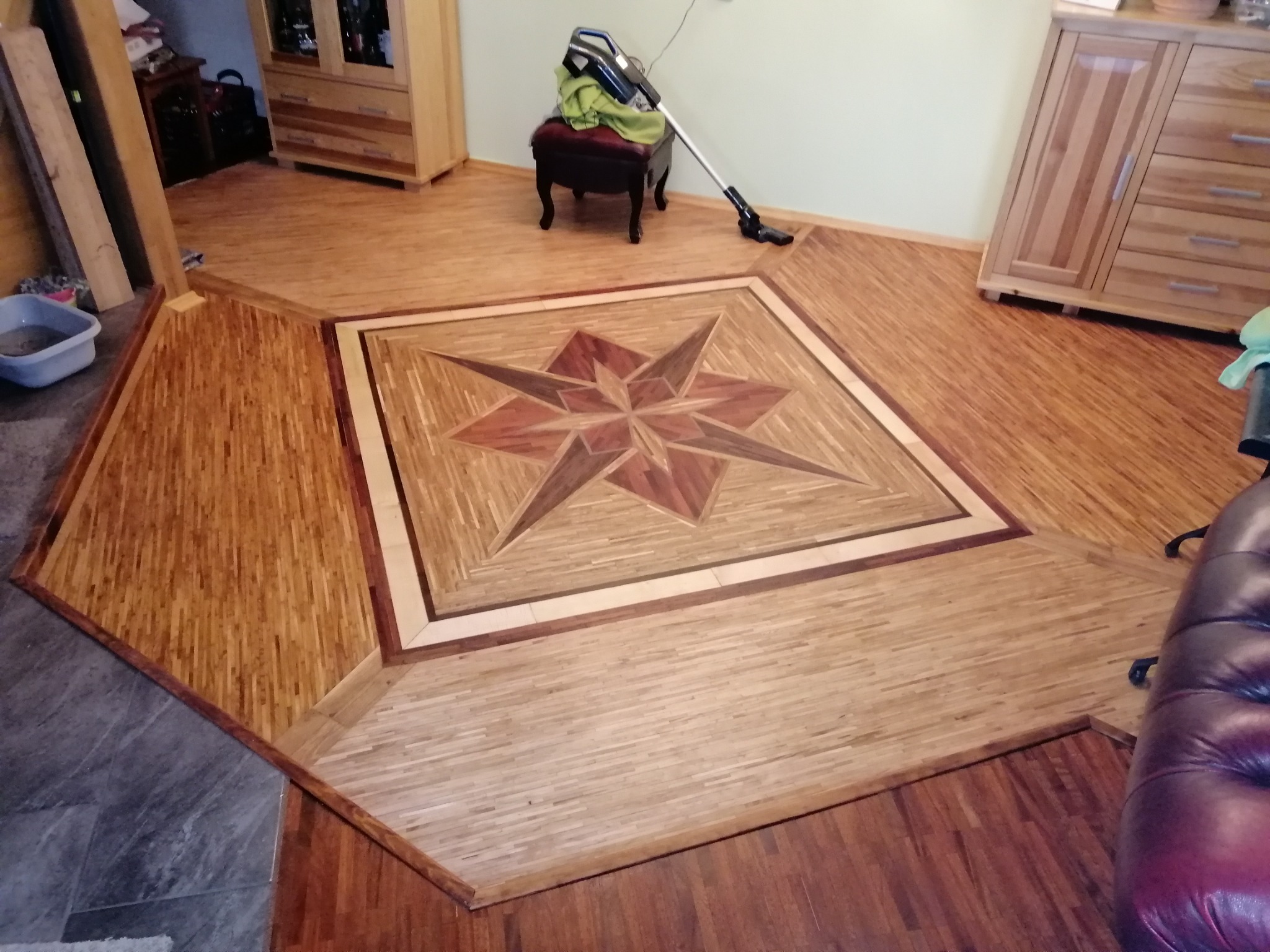Parkiet z drewnianą intarsją w kształcie gwiazdy, otoczony różnymi odcieniami drewna, widoczny odkurzacz na krześle w tle.