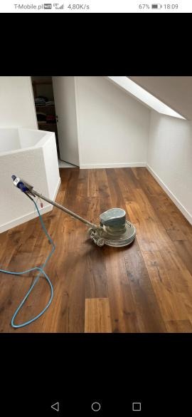 Szlifowanie drewnianej podłogi w pokoju na poddaszu za pomocą szlifierki rotacyjnej, widoczny kabel zasilający i fragment garderoby w tle.