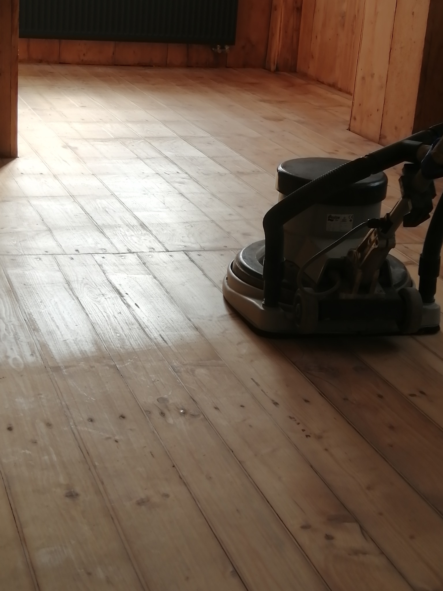 Maszyna do renowacji podłóg drewnianych w trakcie pracy, widoczne świeżo wyszlifowane deski podłogowe i drewniana ściana w tle.