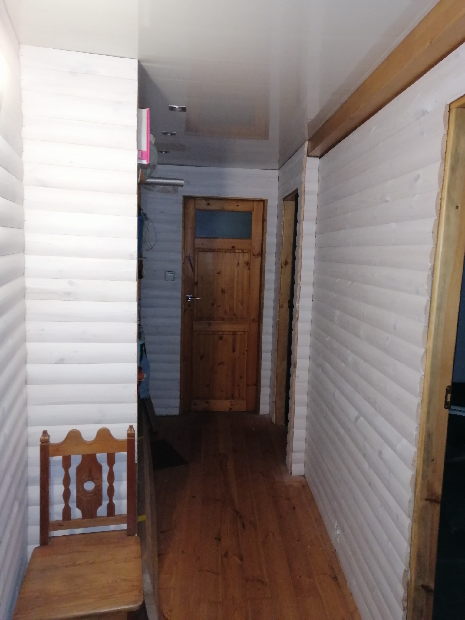 Wąski korytarz wykończony boazerią pomalowaną na biało, z drewnianą podłogą i widokiem na drzwi z szybą oraz drewniane krzesło przy ścianie.