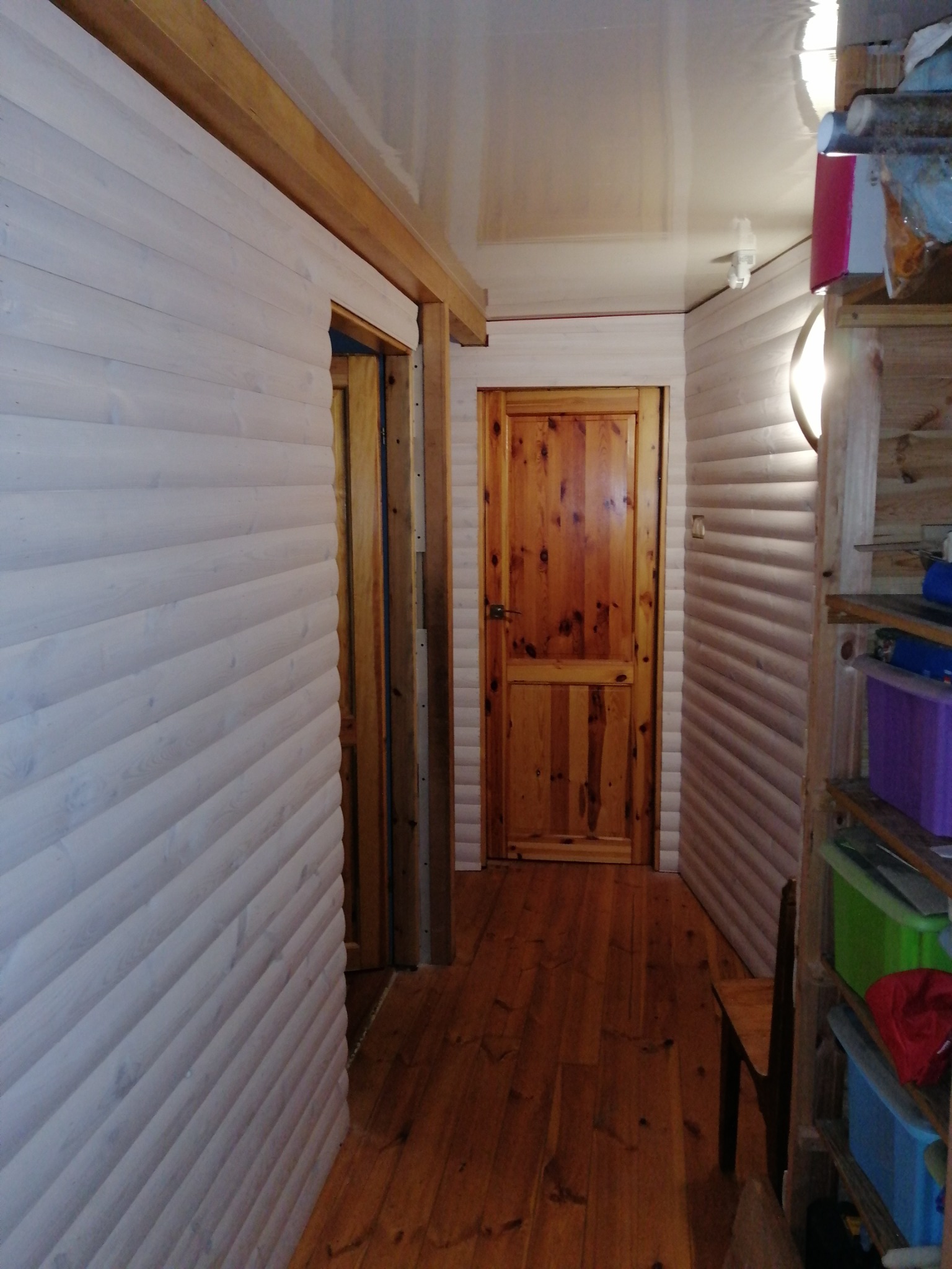 Wnętrze z boazerią pomalowaną na biało i drewnianą podłogą; widoczna otwarta rama drzwiowa i drewniane drzwi w głębi korytarza.